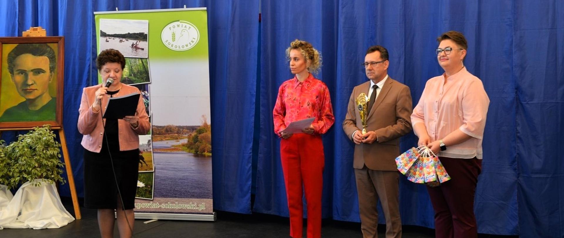 Fotografia wykonana w I LO w Sokołowie Podlaskim podczas rozstrzygnięcia konkursu powiatowego dla szkół podstawowych
pn. „Pokoje zagadek”. Na zdjęciu od lewej dyrektor I LO odczytuje wyniki, z prawej strony stoją kierownik wydziału oświaty i zdrowia Starostwa Powiatowego w Sokołowie Podlaskim, starosta, oraz pracownik I LO. Wszyscy stoją na tle granatowej ściany, roll - upu promocyjnego powiatu sokołowskiego oraz obrazem z wizerunkiem patronki szkoły M. Skłodowskiej - Curie.
