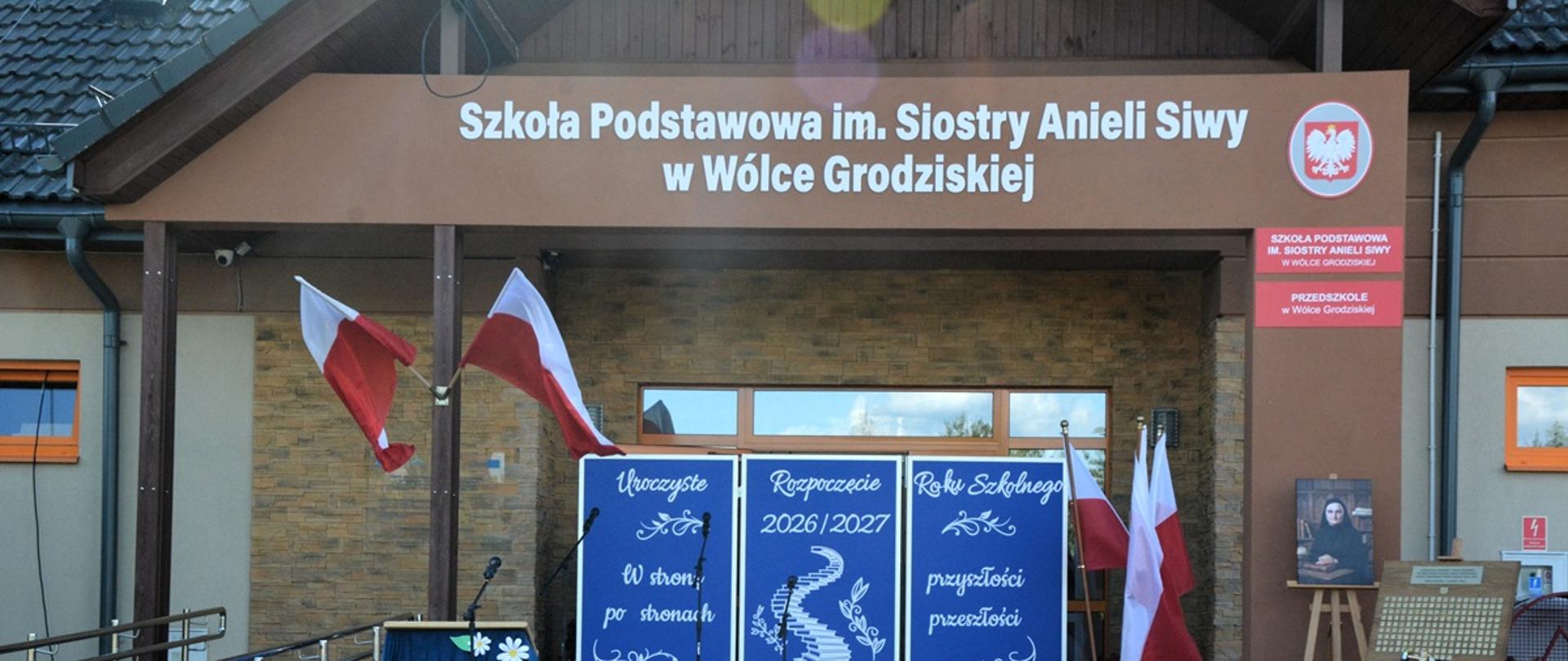 brązowy budynek na górze biały napis szkoła podstawowa imienia siostry anieli siwy w wólce grodziskiej, przed szkoła dekoracja pamiątkowa tablica i zdjęcie siostry w czarnym habicie 