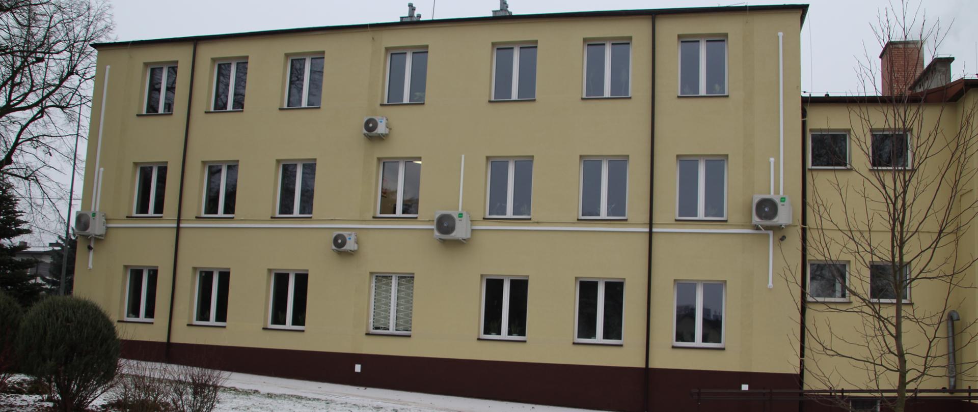 Widok na boczną elewację budynku szkoły w zimowej scenerii. Trawnik przed budynkiem jest lekko przyprószony śniegiem. Na jasnożółtej ścianie zamontowanych jest symetrycznie sześć klimatyzatorów. Po prawej stronie widoczny fragment niższego skrzydła budynku oraz podjazd dla osób z niepełnosprawnościami.