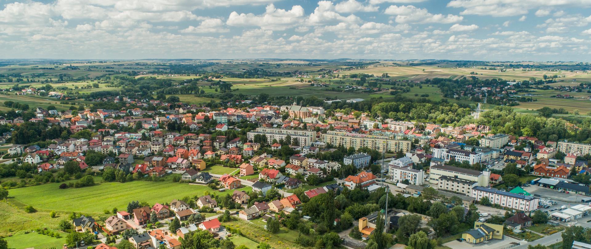 Zdjęcie przedstawia budynki miasta Proszowice z lotu ptaka, w oddali tereny rolnicze Powiatu Proszowickiego.