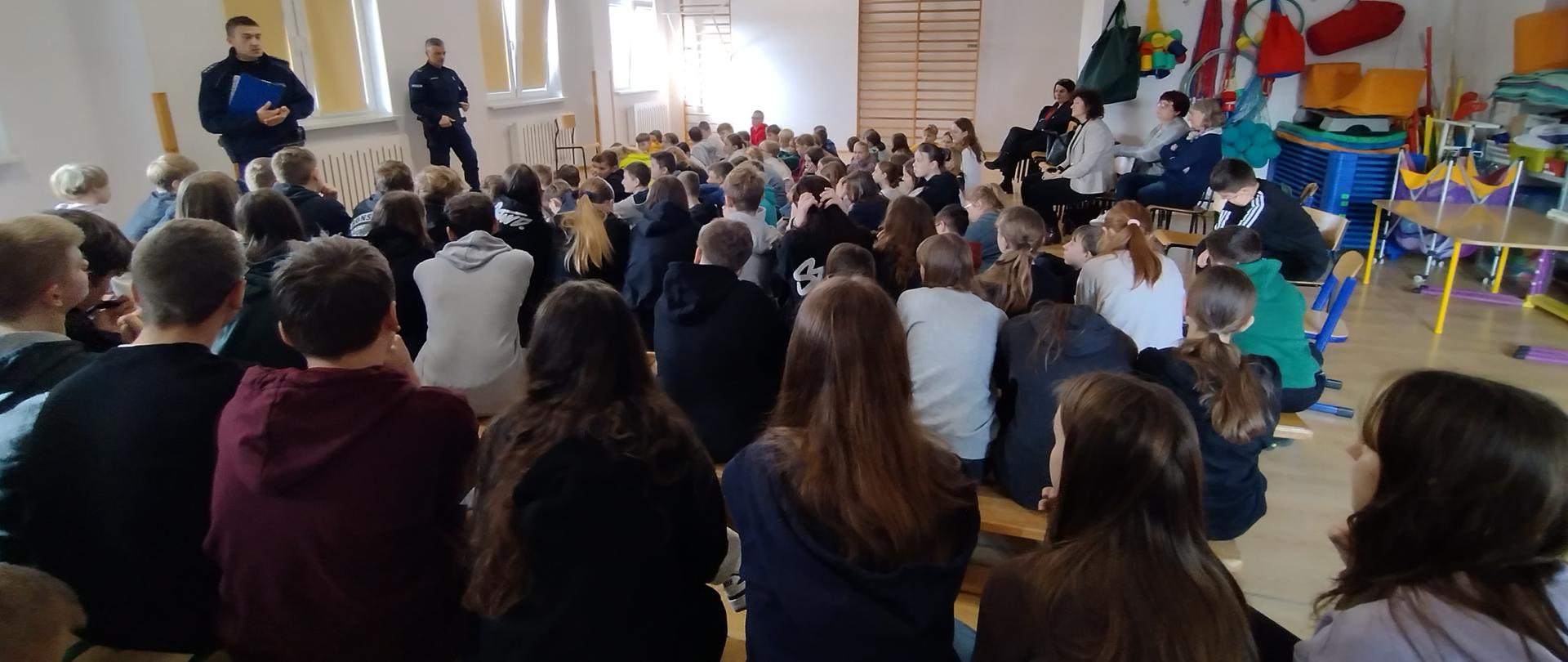 Uczniowie wraz z nauczycielami i dyrekcją siedzący na ławkach i materacach uczestniczą w spotkaniu z dwoma umundurowanymi policjantami.