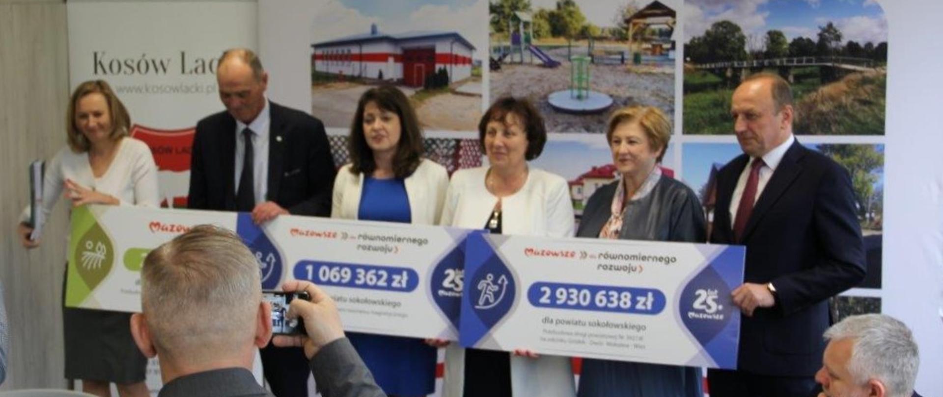 Na zdjęciu członkowie Zarządu Województwa Mazowieckiego wraz z reprezentacją Powiatu Sokołowskiego oraz wójtem Gminy Jabłonna Lacka. Jest to moment wręczenia symbolicznych czeków z dofinansowaniem na realizację trzech zadań inwestycyjnych tożsamych z treścią artykułu. Na czekach wypisane są kwoty dofinansowania oraz nazwa beneficjenta oraz nazwa zadania. W tle ścianka ze zdjęciami z hasłem Mazowsze dla sołectw oraz rollup z napisem Kosów Lacki oraz herbem.