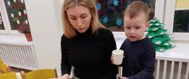 Chłopczyk i jego mama siedzą przy stole, wspólnie tworząc świąteczne ozdoby - papierową torbę z bożonarodzeniowym akcentem