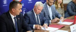 Zdjęcie przedstawia moment podpisania umowy na budowę drogi. Na zdjęciu przedstawiciele Powiatu Kraśnickiego, firmy Wod-Bud i Zarządu Dróg Powiatowych w Kraśniku siedzą za stołem. Na stole znajdują się egzemplarze umów, na których Starosta Kraśnicki oraz przedstawiciel firmy Wod-Bud składają podpisy. W tle znajduje się niebieska ścianka Powiatu Kraśnickiego z wizerunkiem herbu powiatu.