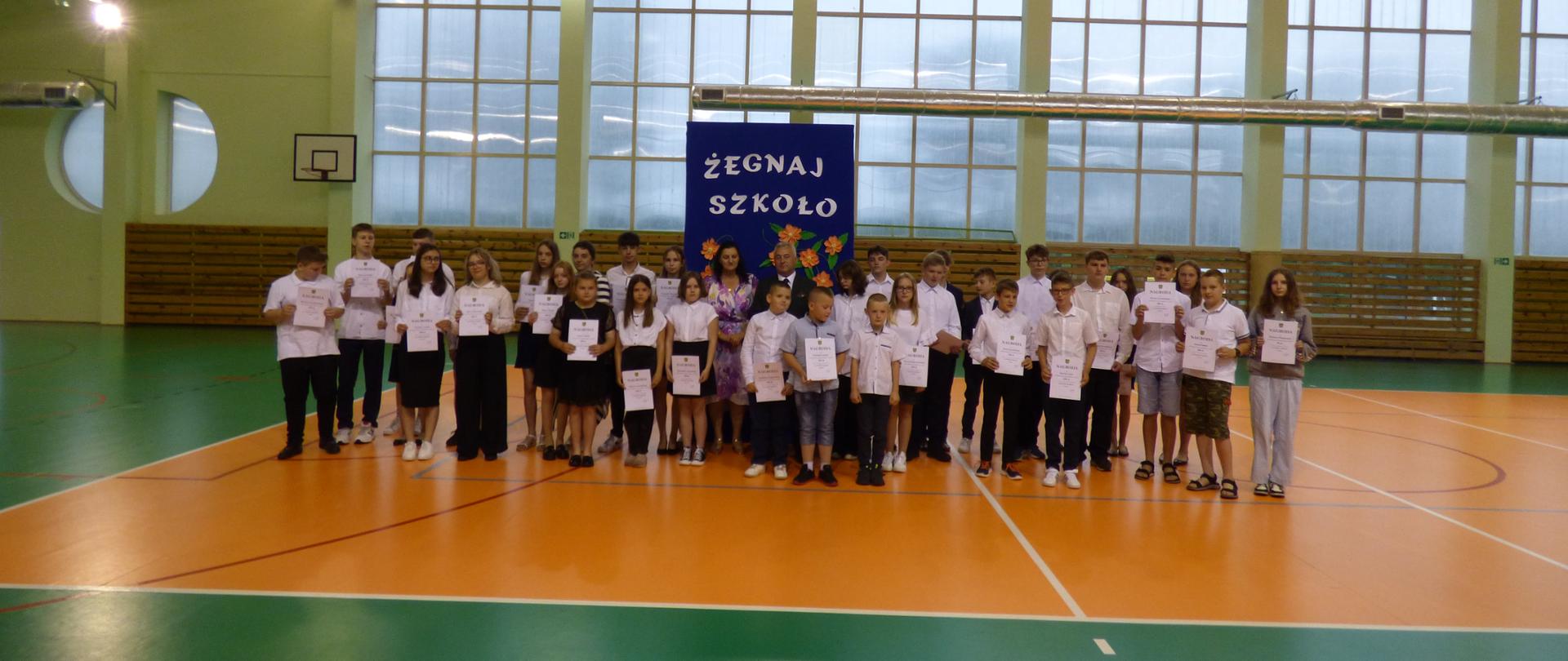 Wszyscy nagrodzeni stypendiami stoją na tle niebieskiej tablicy z napisem ŻEGNAJ SZKOŁO, trzymają w rękach dyplomy. W tle widać halę gimnastyczną. 