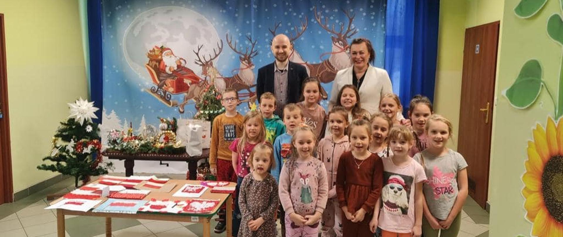 Pani Wójt Gminy Łabowa Marta Słaby oraz Pan Dawid Madziar, Prezes Fundacji Socius, wręczają uczniom klas 1-3 Szkoły Filialnej w Roztoce Wielkiej nagrody za udział w konkursie plastycznym w ramach projektu "W sercu Ojczyzny", na stoliku leżą prace plastyczne uczniów, w tle dekoracja ze świętym Mikołajem, po prawej stronie uczniowie odbierający nagrody