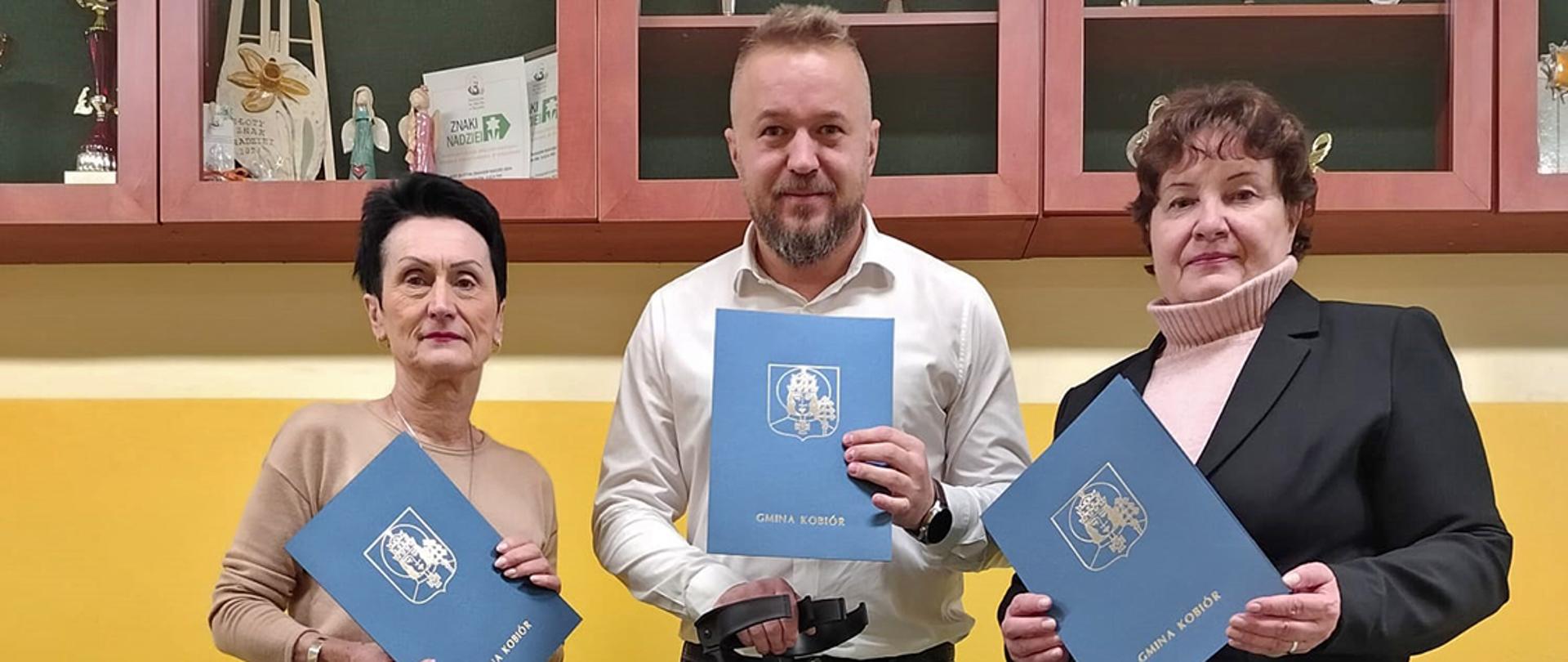 Trzy osoby trzymające niebieskie ozdobne etui na tle żółtej ściany