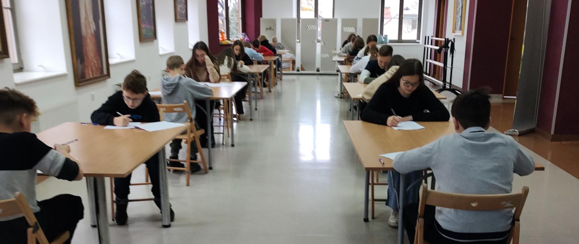 Na dużej sali uczniowie siedzą przy stolikach i rozwiązują pisemny test 