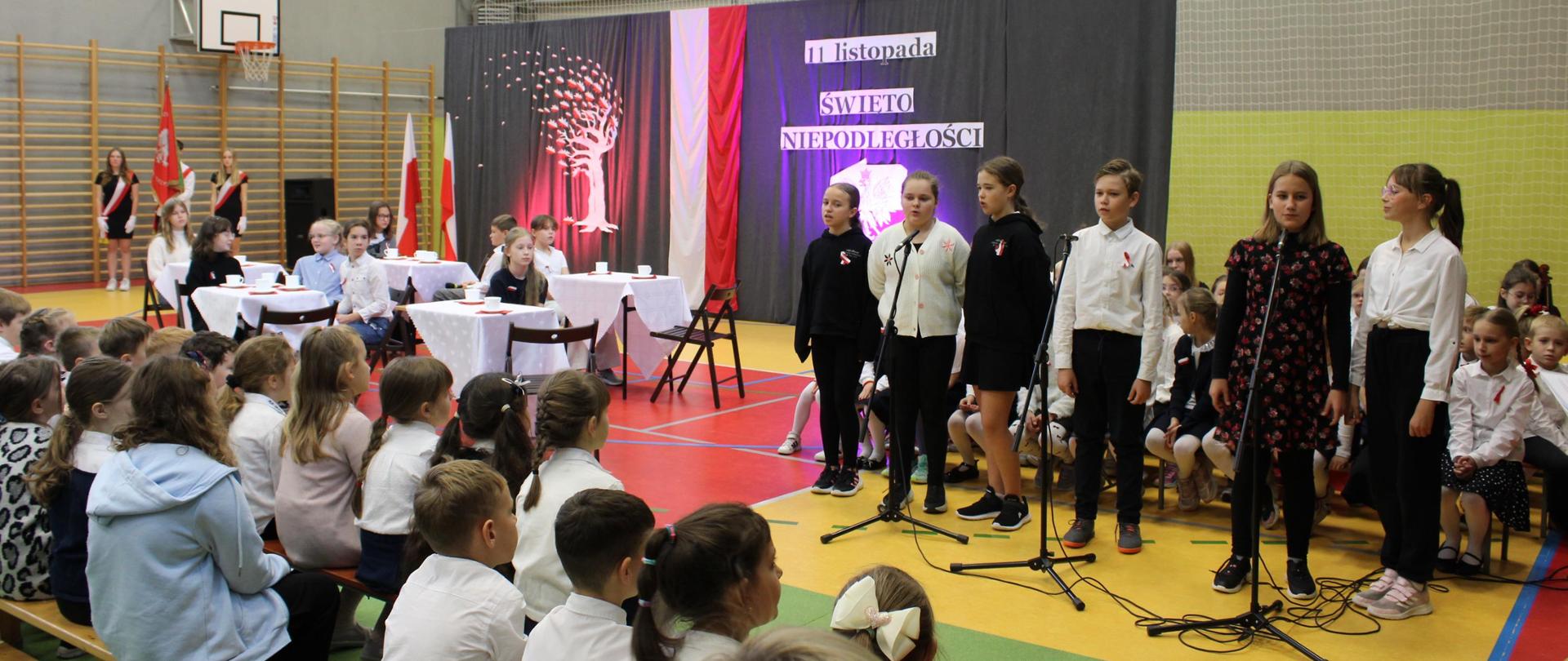 Koncert uczniów na sali gimnastycznej. Uczniowie stoją przed mikrofonami. Część siedzi przy stolikach. Reszta uczniów patrzy na wytęp siedząc na ławkach.