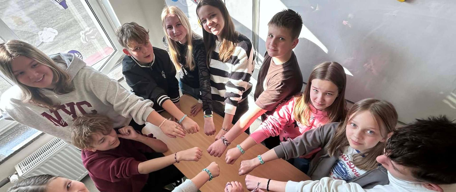 Grupa dzieci stoi w kółeczku z wystawioną ręką do środka na której są kolorowe bransoletki.