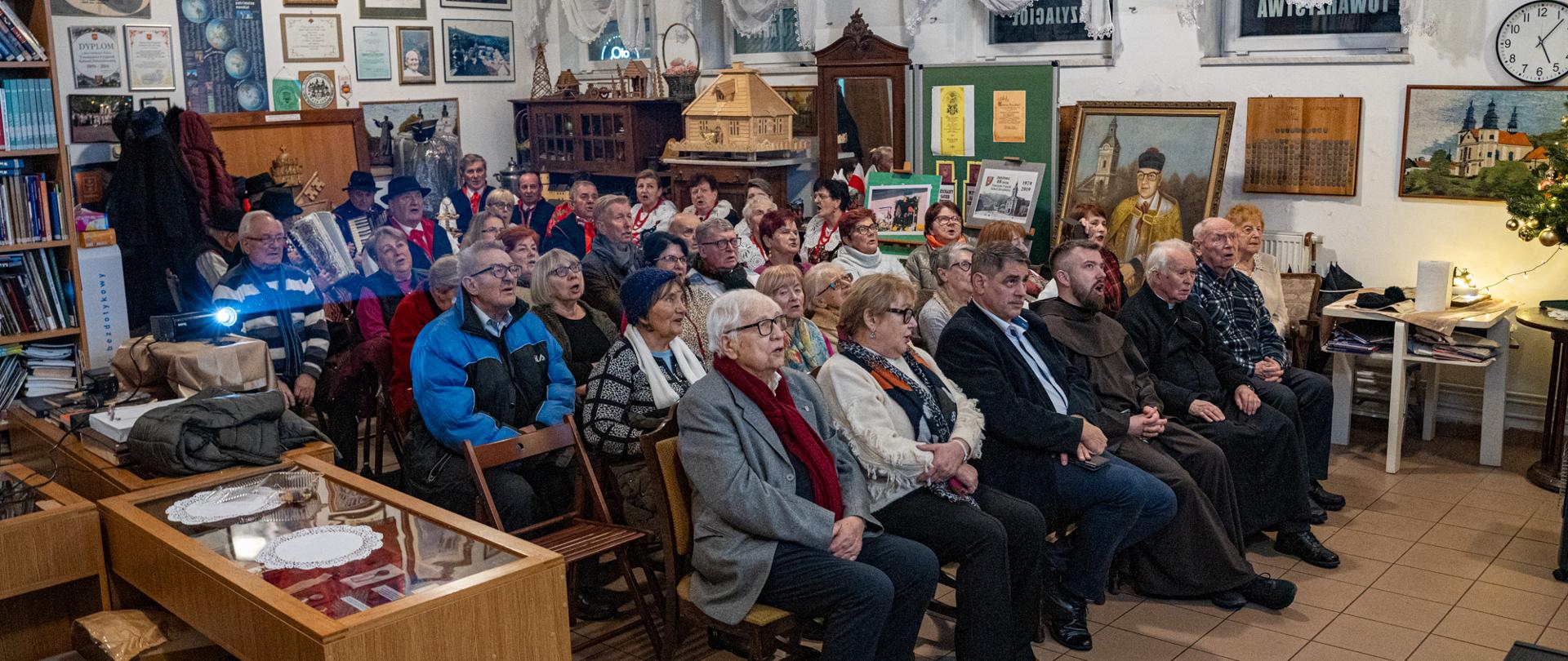 Grupa kilkudziesięciu osób, głównie seniorów, siedzi na krzesłach w niewielkiej sali pełnej pamiątek, obrazów i sztandarów. Uczestnicy wspólnie śpiewają kolędy, patrząc w jednym kierunku. W tle widoczne są gabloty muzealne, portrety oraz dekoracje świąteczne, a po lewej stronie projektor wyświetlający obraz.
