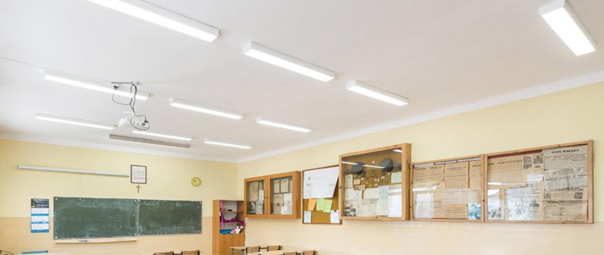 Lampy ledowe w sali lekcyjnej na białym suficie. Poniżej tablica gabloty i ławki oraz krzesła.
