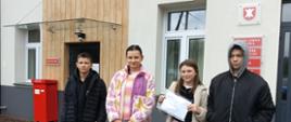 Dwie uczennice i dwóch uczniów stoją przed urzędem pocztowym w Łabowej, jedna uczennica trzyma w ręku list, za nimi widoczna czerwona skrzynka na listy