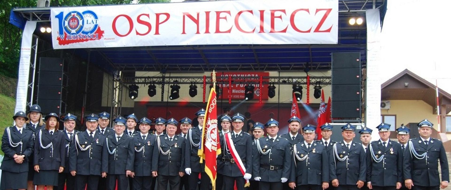 Zdjęcie grupowe Strażaków z Jednostki OSP w Niecieczy ze Sztandarem jednostki. Strażacy ustawieni w dwuszeregu. W tle scena, a na jej szczycie biały baner z logo 100 lat w jedności i napisem OSP Nieciecz.