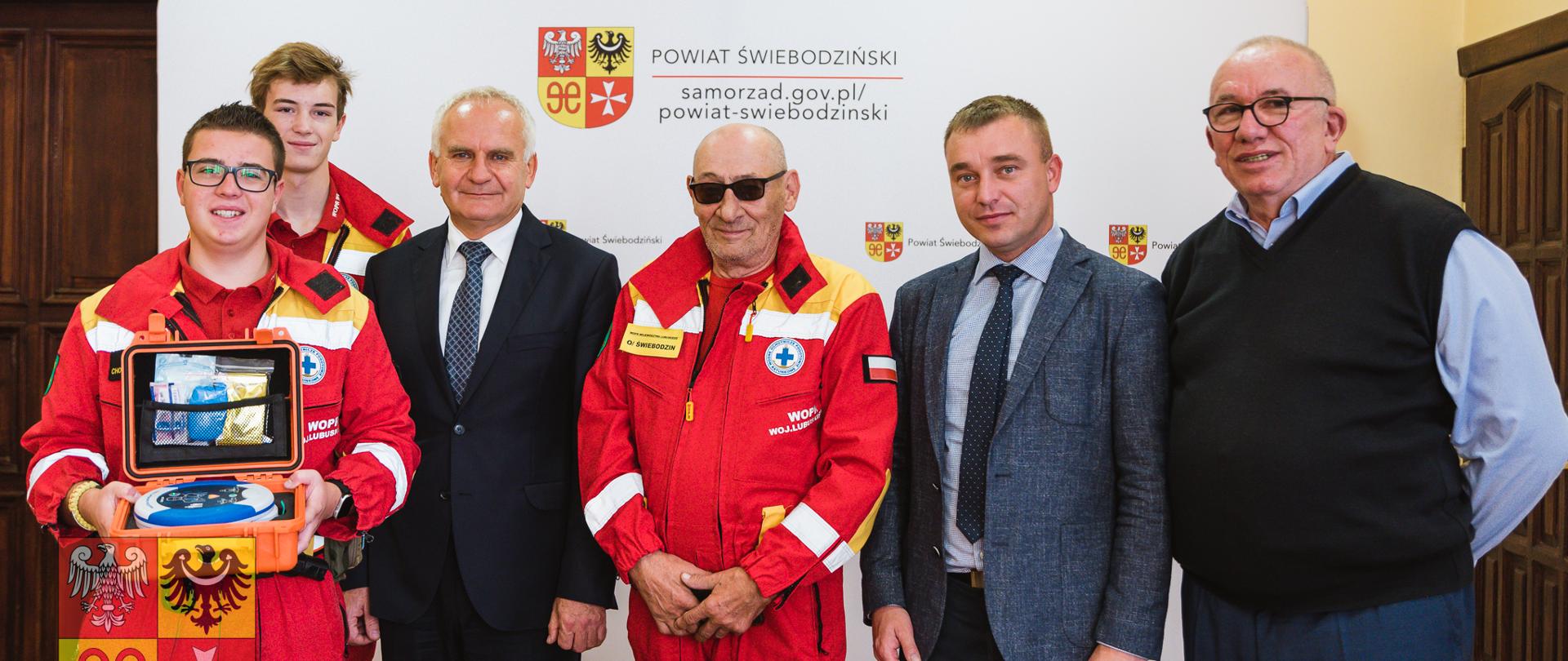 grupa mężczyzna wewnątrz, trzej w czerwonym kombinezonie ratownika, jeden trzyma defibrylator