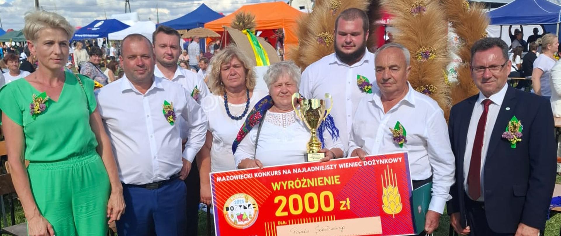 Na zdjęciu członkowie Delegacji Wieńcowej wraz ze starostą, trzymający Voucher nagrodę oraz puchar. W tle wieniec Dożynkowy.