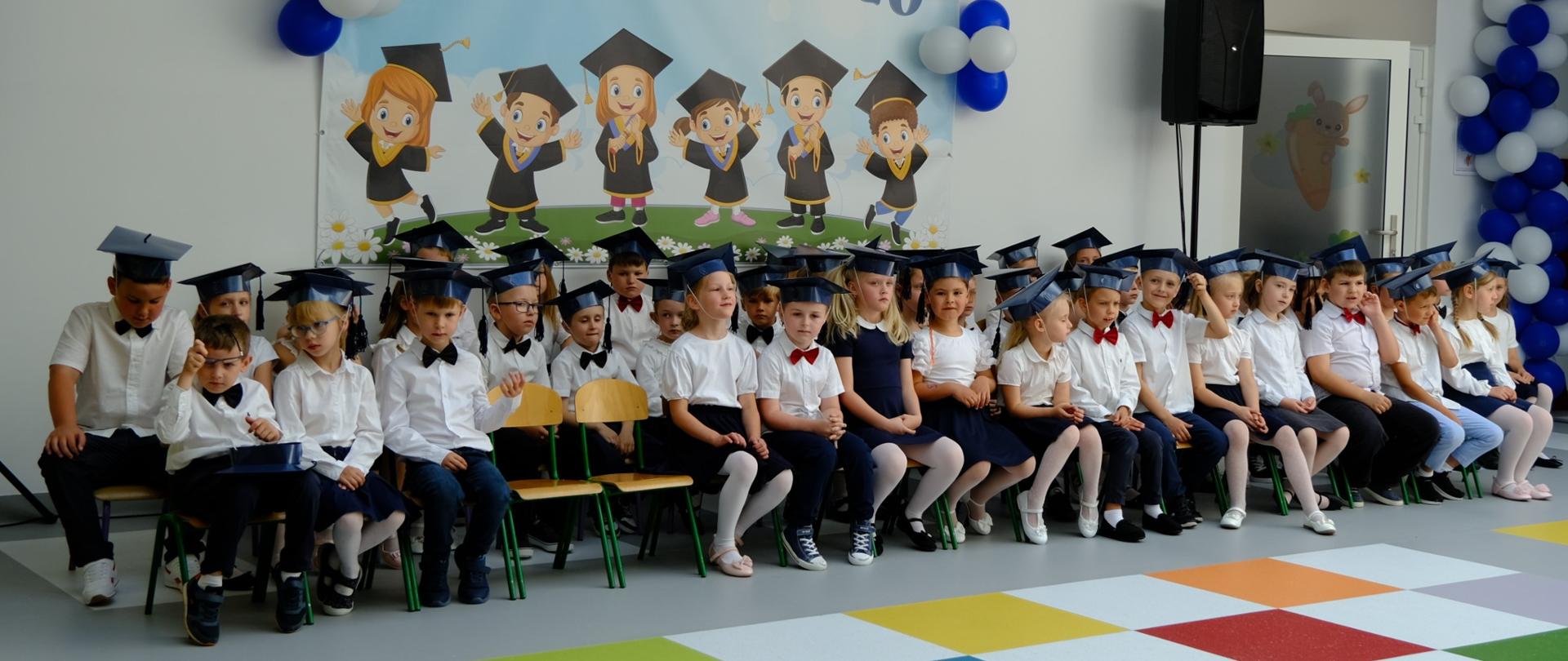 Grupa dzieci w biało-granatowych strojach siedzi w białej sali