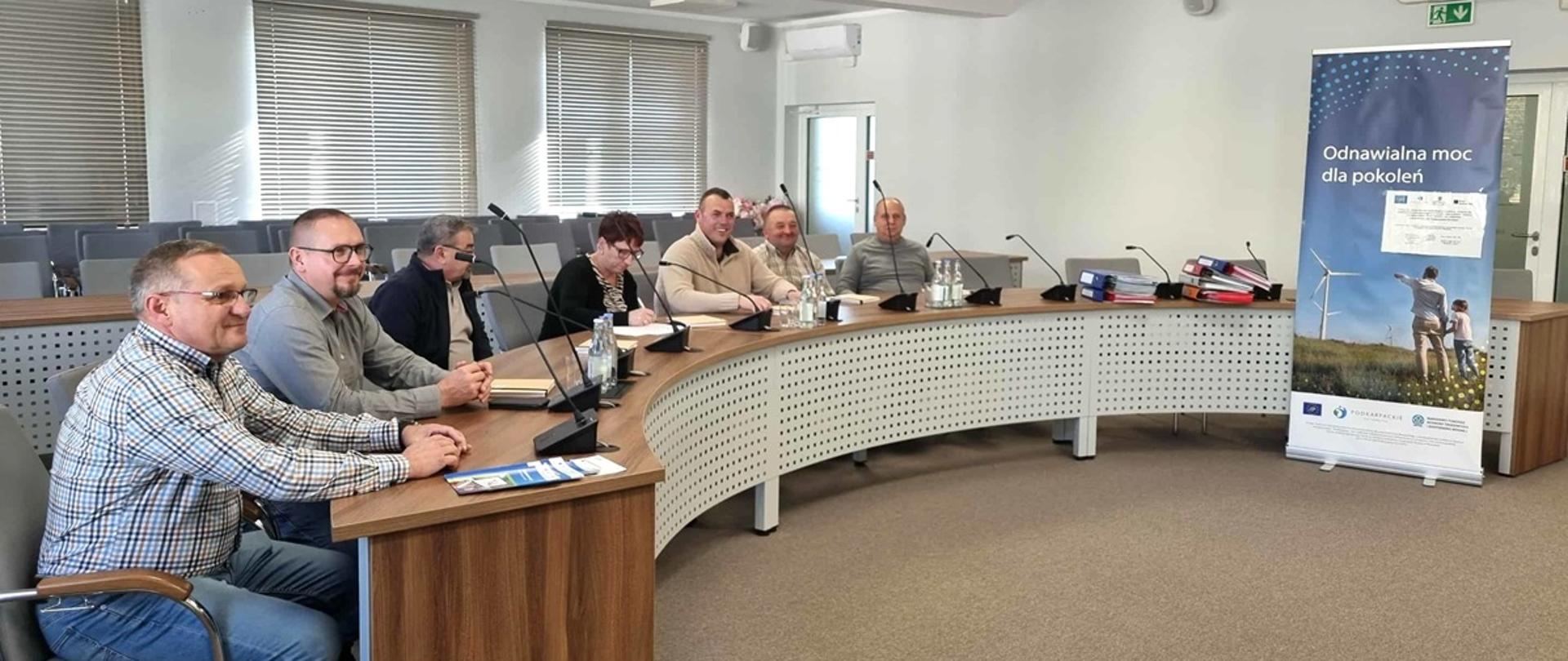 sześciu mężczyzn i jedna kobieta siedzą za półokrągłym stołem na sali konferencyjnej 