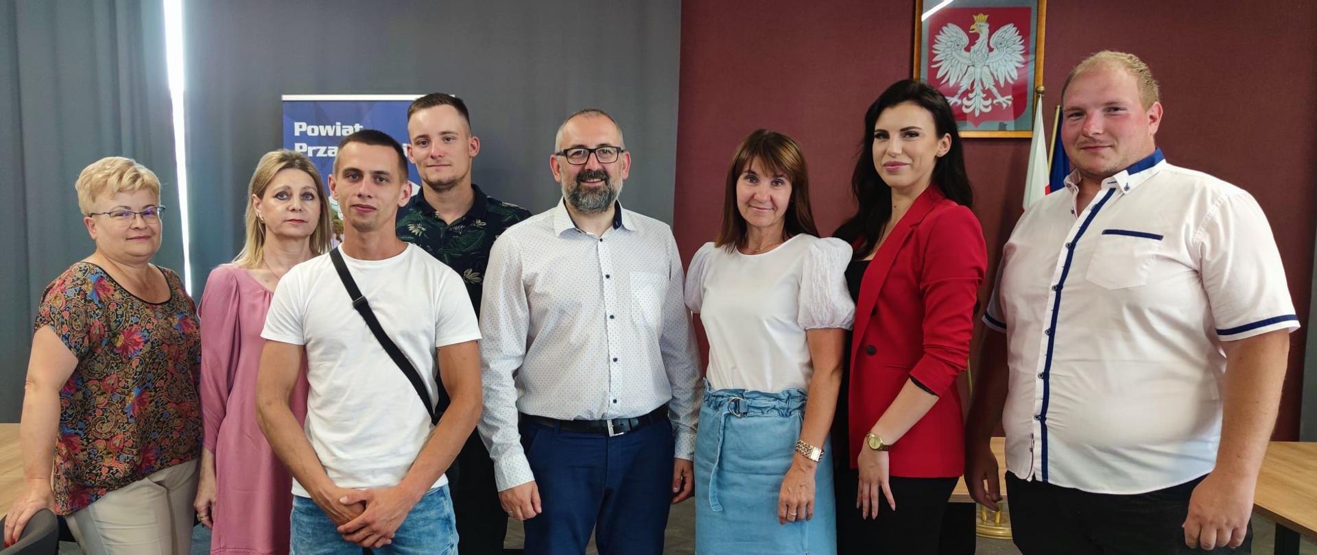 Zdjęcie przedstawia Starostę Przasnyskiego Krzysztofa Bieńkowskiego, Wicedyrektor Powiatowego Urzędu Pracy Annę Obrębską oraz Panią Barbarę Garlińską wraz z nowymi przedsiębiorcami powiatu przasnyskiego tuż po podpisaniu umów na dofinansowanie rozpoczęcia ich działalności gospodarczej. Zdjęcie wykonano na sali konferencyjnej Centrum Aktywizacji Przedsiębiorczości w Starostwie Powiatowym w Przasnyszu, w tle szare kotary, ściany w kolorze szlachetnego mahoniu oraz oprawione godło Rzeczypospolitej Polskiej – biały orzeł w złotej koronie na czerwonym tle.