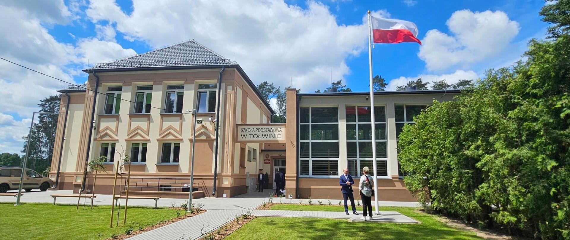 Budynek z polską flagą przed wejściem, otoczony zielenią i przechodniami.
