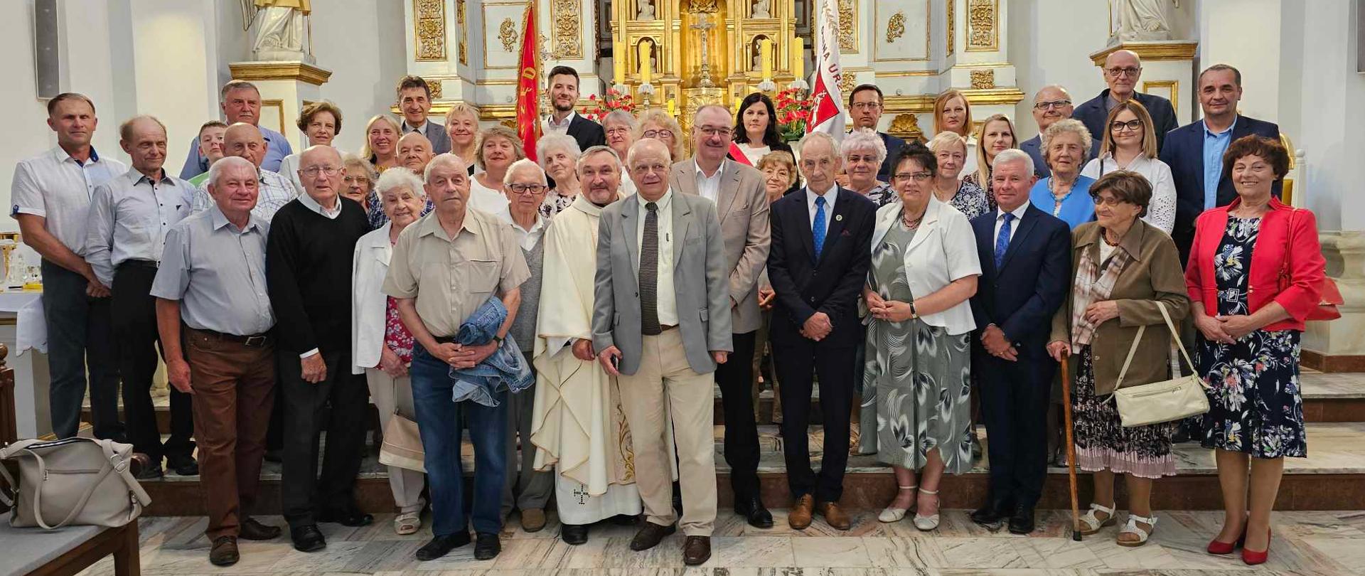 Pamiątkowe zdjęcie członków Towarzystwa Ziemi Urzędowskiej przedstawia grupę ludzi zebranych na stopniach przed ołtarzem. Pozłacany ołtarz znajdujący się w tle ozdobiony jest złotymi kolumnami, figurami świętych i ornamentami.