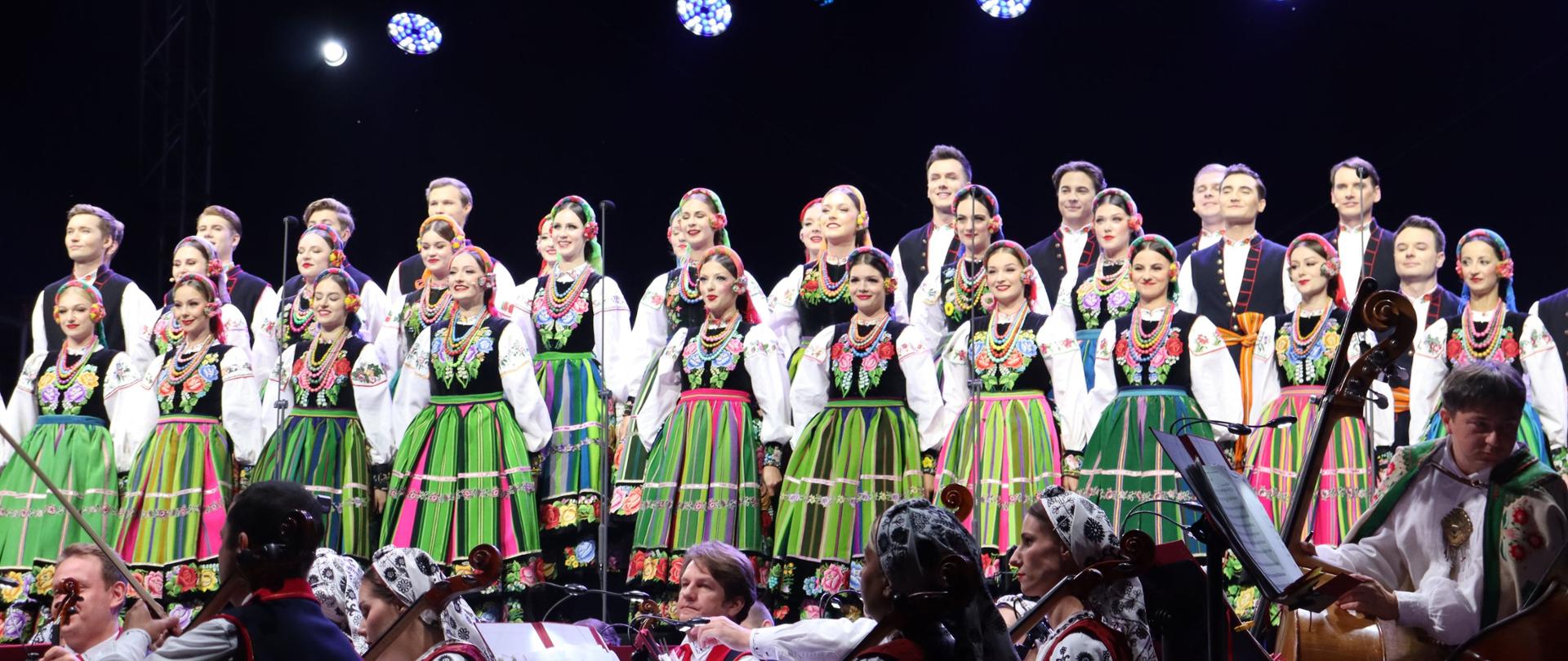 Duża grupa artystów ustawiona na scenie podczas wspólnego występu. Kobiety prezentują charakterystyczne pasiaste, kwieciste stroje, a za nimi stoją mężczyźni. Na pierwszym planie widoczna jest orkiestra.