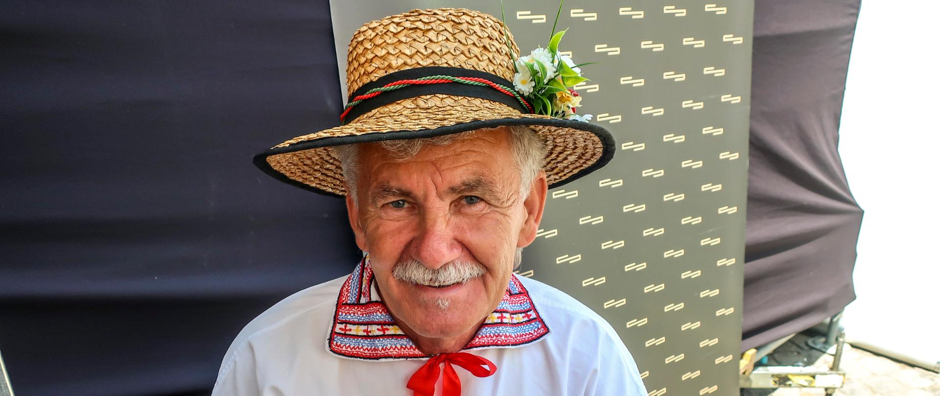 Zdjęcie przedstawia starszego mężczyznę w białej koszuli ludowej z kolorowym, haftowanym kołnierzem i czerwoną kokardką. Na głowie ma słomiany kapelusz.