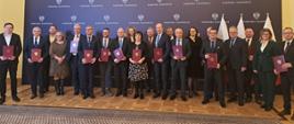 Zdjęcie grupowe, na którym widzimy przedstawicieli samorządów wraz z przedstawicielami Mazowieckiego Urzędu Wojewódzkiego w Warszawie. Za grupą stojących w dwóch rzędach osób widoczna ciemna (granatowa) ścianka z białymi logotypami Wojewody Mazowieckiego składającym się z białego orła w koronie oraz podpisu Wojewoda Mazowiecki. Logotypy ułożone w rządkach od góry do dołu ścianki.