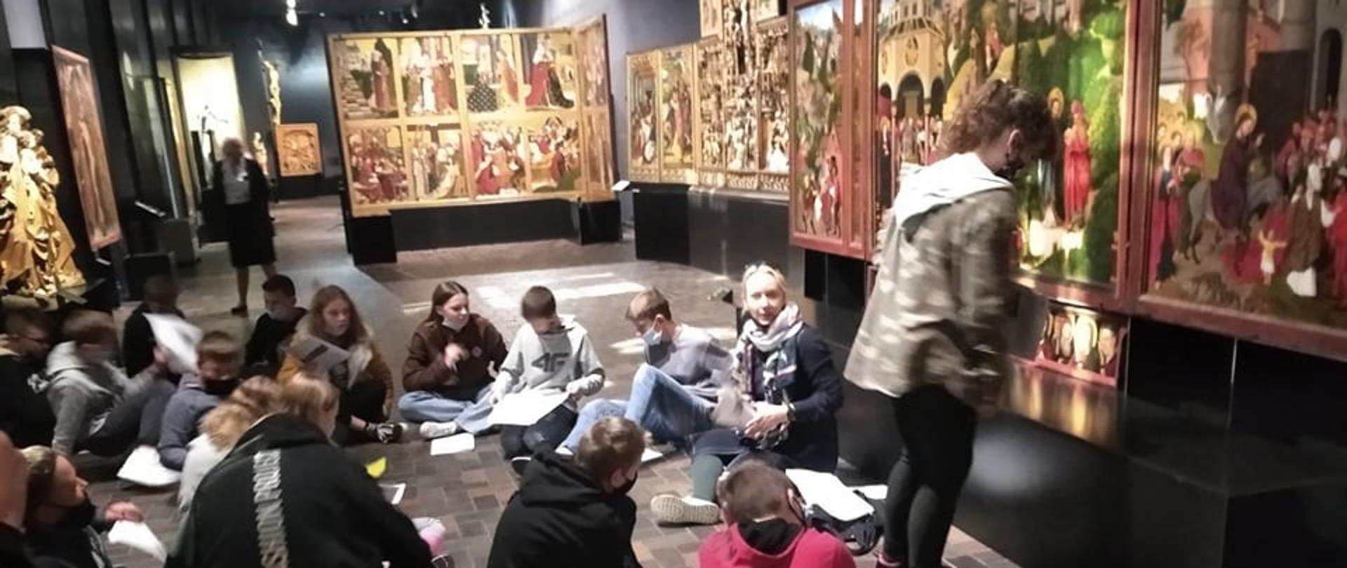 Młodzież siedzi na podłodze i trzyma broszury. Na ścianach wiszą obrazy
