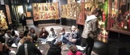 Młodzież siedzi na podłodze i trzyma broszury. Na ścianach wiszą obrazy