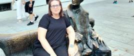 Uczestniczka wycieczki pozuje do zdjęcia na ławce z pomnikową postacią historyczną