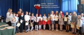 Dzieci ustawione wokół sali. Na ścianie niebieski materiał a na nim biało czerwone kwiaty