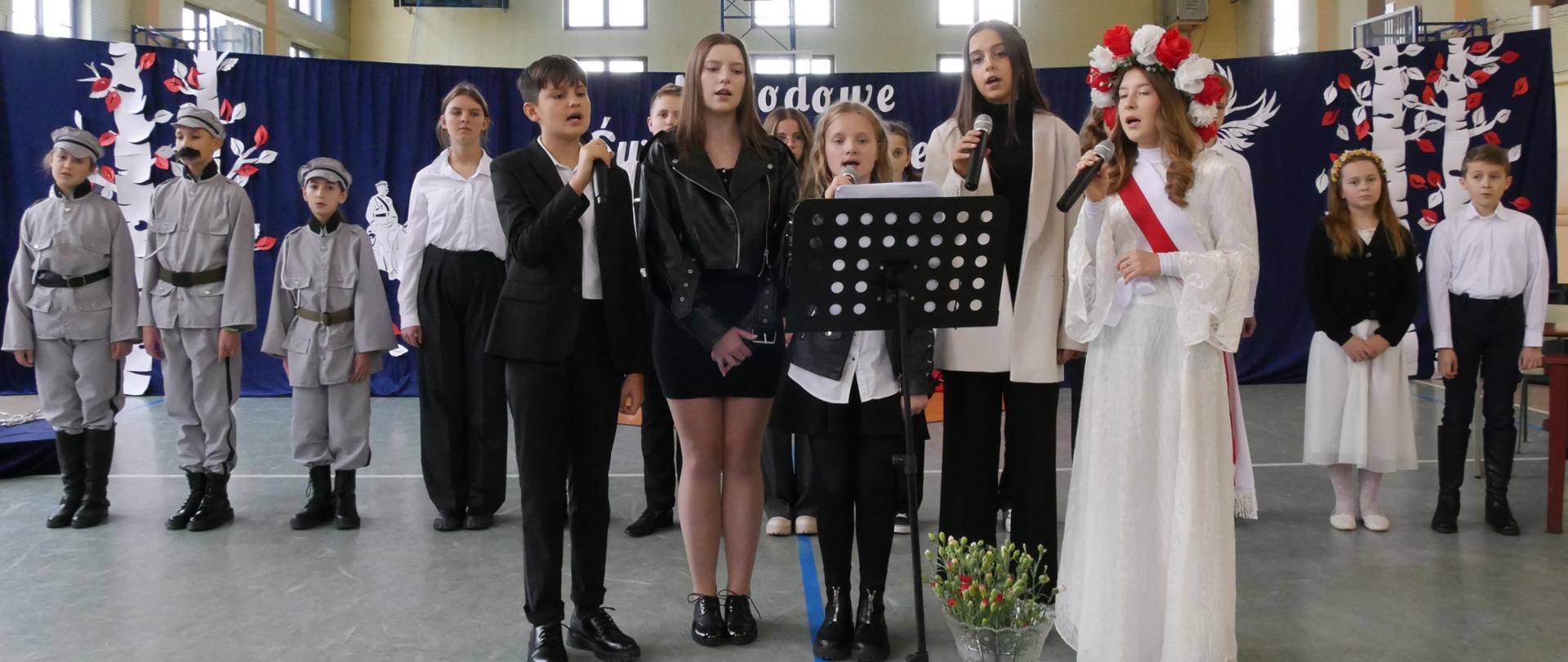 Uczniowie w odświętnych strojach przygotowanych na tle dekoracji w barwach narodowych prezentującej brzozy, Piłsudskiego oraz orła rozdzielającego szponami biało- czerwoną flagę polski pod napisem "Narodowe Święto Niepodległości".