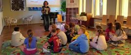 Dzieci siedzą na dywanie i słuchają pani pracującej w bibliotece.