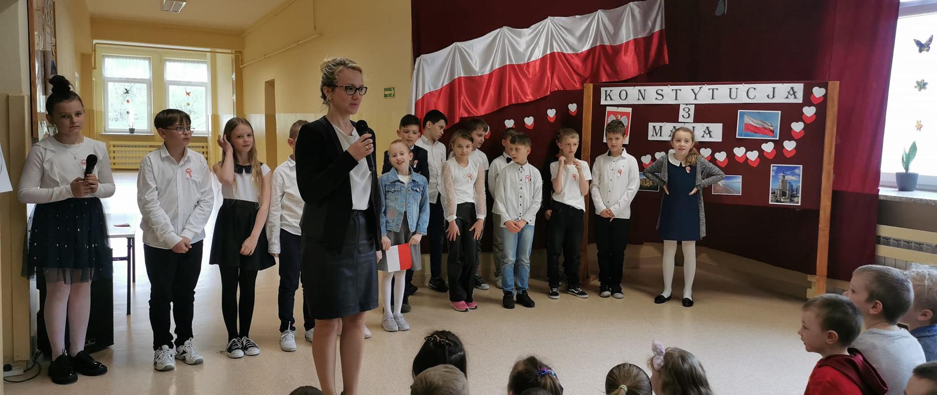 Dyrektor szkoły trzyma w ręku mikrofon, w tle dzieci występujące w akademii z okazji 232. rocznicy uchwalenia Konstytucji 3 Maja, biało czerwona flaga i dekoracje