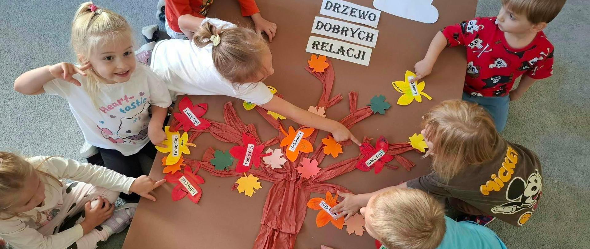 Na zdjęciu widać grupę dzieci wspólnie tworzących kolorową pracę plastyczną przedstawiającą „Drzewo Dobrych Relacji”. 🌳❤️
Dzieci przyklejają do papierowego drzewa kolorowe liście i kwiaty z hasłami, m.in. „Lubię Cię!”. Wspólna praca staje się okazją do rozmowy o życzliwości, przyjaźni, dobrych relacjach i wzajemnym szacunku. Kolorowa kompozycja pokazuje, że dobre słowa i gesty mogą tworzyć atmosferę bezpieczeństwa i wspólnoty. 🤝🌈