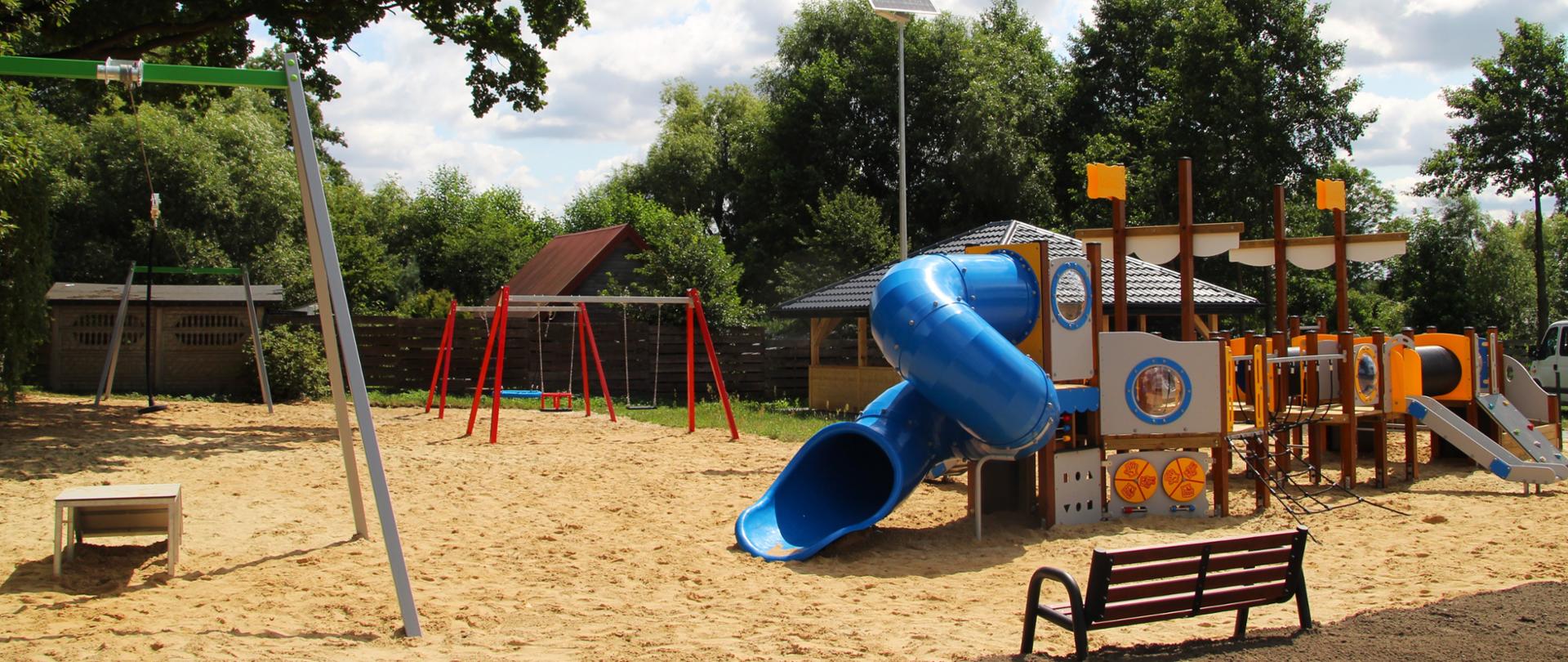 Plac zabaw o piaszczystym podłożu otoczony wysokimi drzewami. Na pierwszym planie widoczny jest wielofunkcyjny zestaw zabawowy w formie statku z dużą, niebieską zjeżdżalnią rurową oraz drewniana ławka. W tle znajdują się klasyczne huśtawki oraz zjazd linowy.