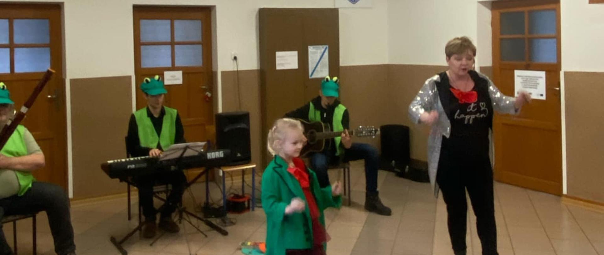 Na scenie stoi jasnowłosa dziewczynka ubrana w czerwoną sukienkę, zielony frak i czerwoną muszkę. Obok niej stoi Pani ubrana w czarną koszulkę i czarne spodnie. Na sobie ma również srebrny frak i czerwoną muszkę. Za nimi, na krzesłach siedzi troje muzyków. Mężczyźni grają na instrumentach, m.in. keybordzie i gitarze. Wszyscy ubrani są w zielone kamizelki i zielone czapki.