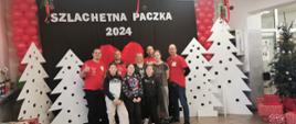 Grupa 9 osób stoi na tle ścianki z napisem Szlachetna paczka o dekoracji w postaci białych choinek oraz serca