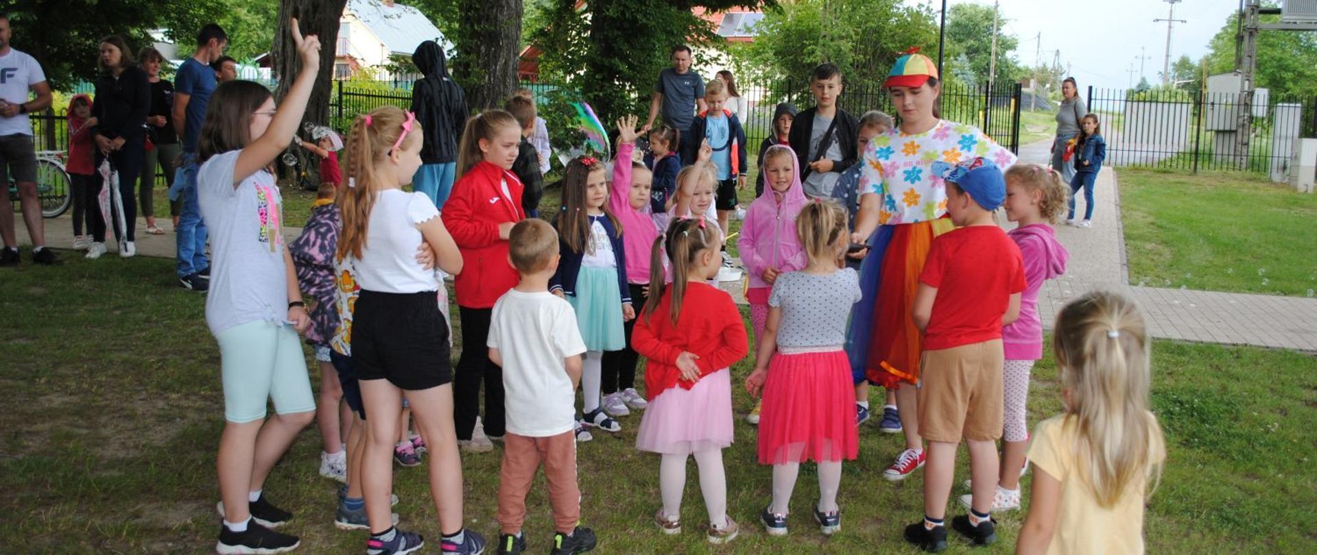 dzieci kolorowo ubrane stoją w grupie, pani w kolorowej spódnicy trzyma mikrofon do którego dzieci mówią 