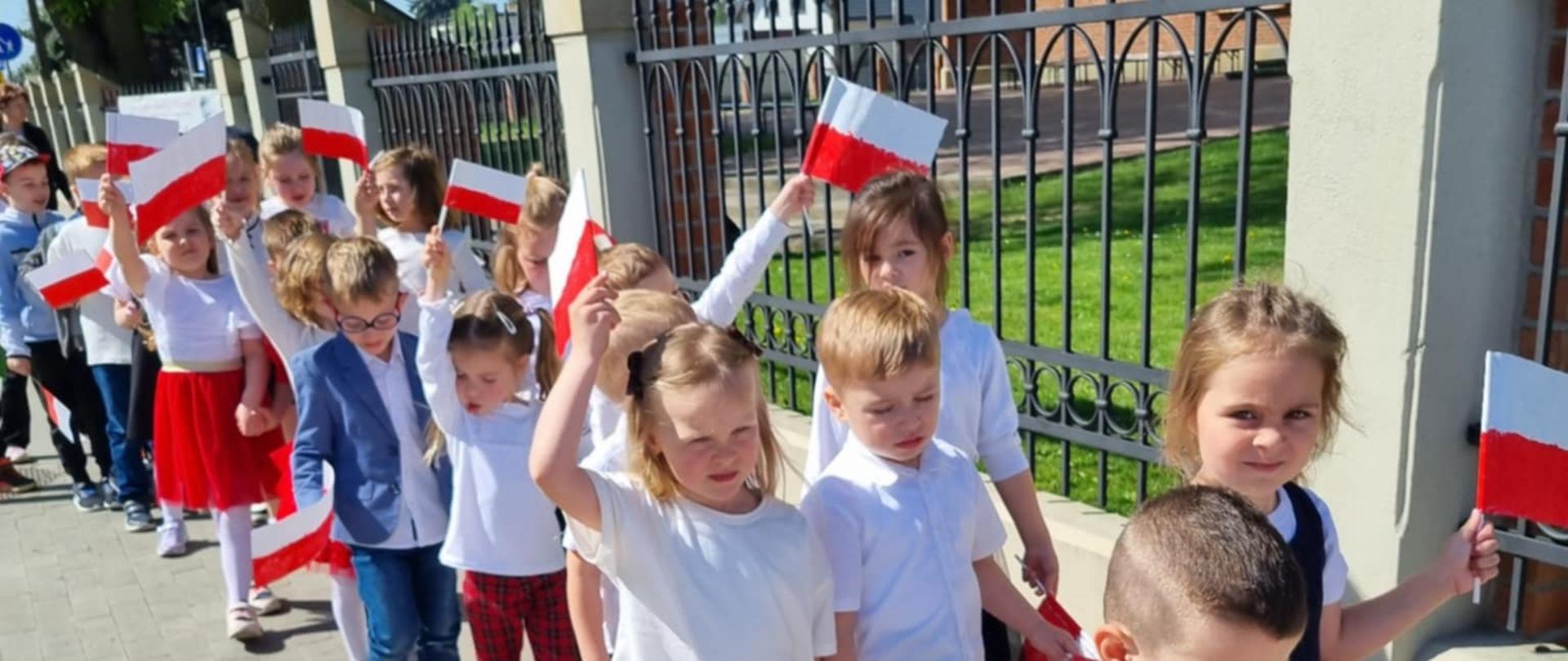 Grupa dzieci spaceruje ulicami Chorzelowa. Dzieci ubrane w barwy narodowe, trzymają w rękach flagi Polski.