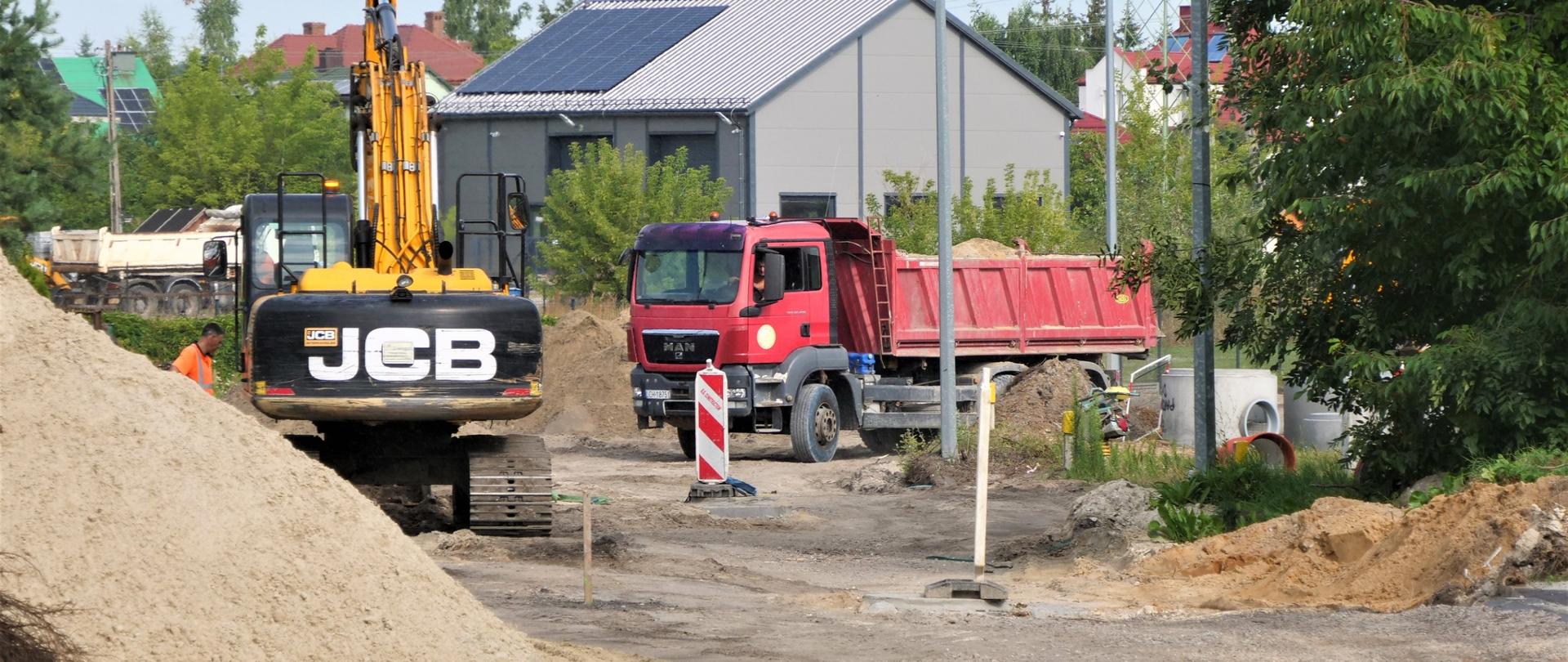Zdjęcie przedstawia prace budowlane na drodze. Na rozkopanym odcinku drogi stoi koparka oraz ciężarówka