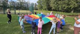 Grupa dzieci bawiących się na świeżym powietrzu, trzymają i unoszą kolorową chustę do animacji. W tle boisko i drzewa.