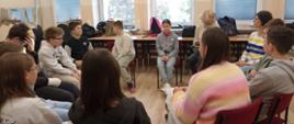 Uczniowie siedzą na krzesłach ustawionych w kole w swojej klasie, między nimi siedzi pani psycholog prowadząca zajęcia i nauczycielka języka polskiego