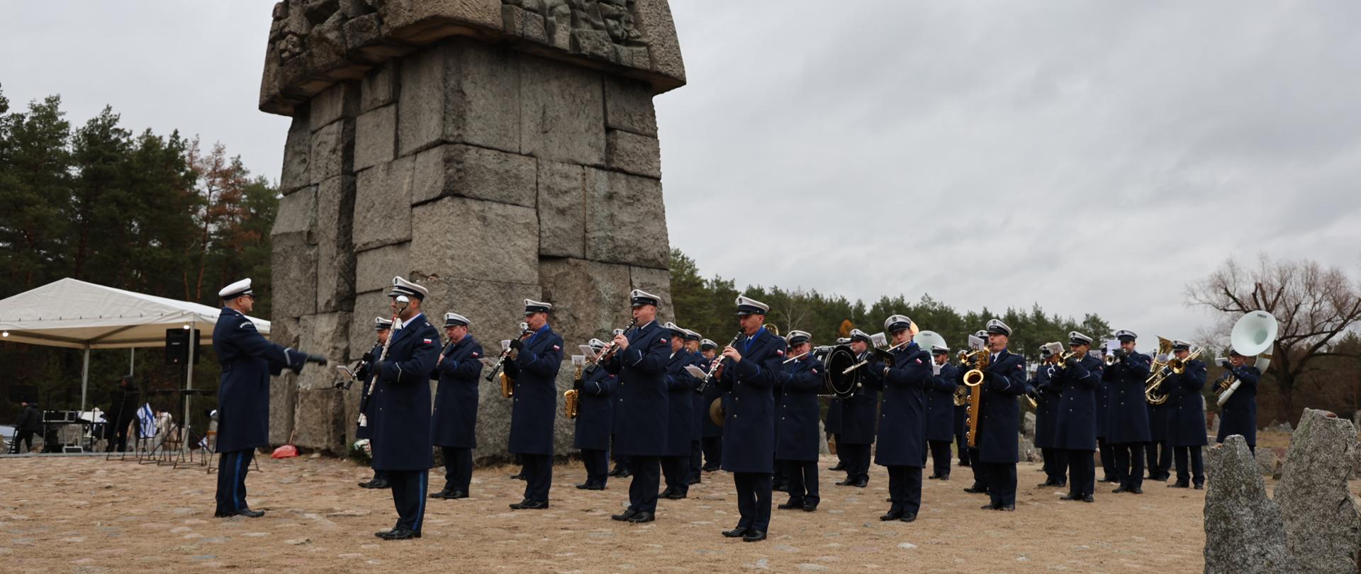 Na tle pomnika głównego widzimy stojącą orkiestrę reprezentacyjną Komendy Głównej Policji. Policjanci w mundurach galowych, z białymi czapkami na głowie grają pod pomnikiem na instrumentach dętych. Przed nimi dyrygent. W oddali widzimy ścianę lasu.
