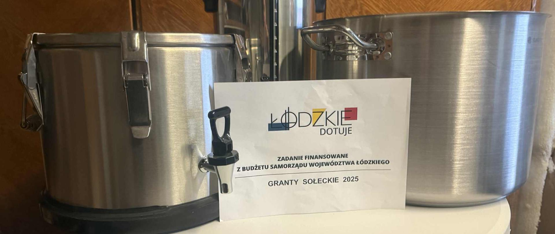 Sprzęt gastronomiczny ze stali nierdzewnej: termos z kranikiem, duży garnek oraz warnik do wody. Przed przedmiotami stoi biała kartka z logo „Łódzkie dotuje” i napisem: „Zadanie finansowane z budżetu Samorządu Województwa Łódzkiego – Granty Sołeckie 2025”.