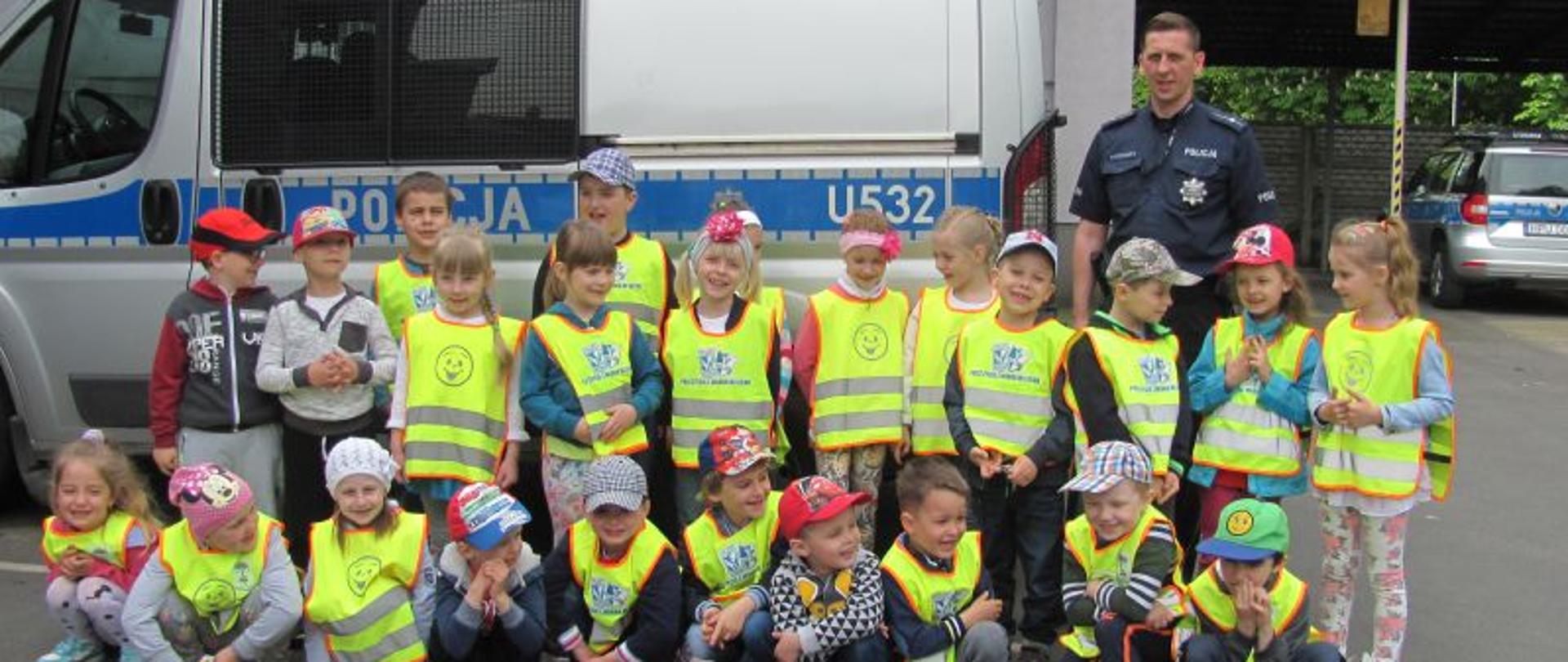 Grupa dzieci w kamizelkach odblaskowym stoją lub kucają przed radiowozem policyjnym. Za dziećmi stoi policjant.
