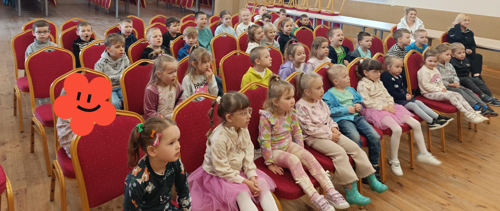 Zdjęcie przedstawia dużą grupę dzieci w wieku przedszkolnym, które siedzą na dużych czerwonych krzesłach ustawionych w rzędach na dużej sali. Obok dzieci siedzą dwie dorosłe kobiety. Po prawej stronie okno i drzwi. W tle widać stoły na których poukładane są krzesła.