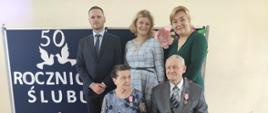 Państwo Henryka i Adam na tle granatowej tablicy z napisem wraz z Wójtem, Z-cą Wójta i Z-cą Kierownika USC