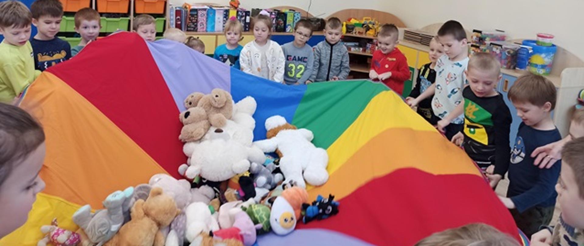 Dzieci trzymają chustę animacyjną, na której huśtają wszystkie misie przyniesione do przedszkola. 